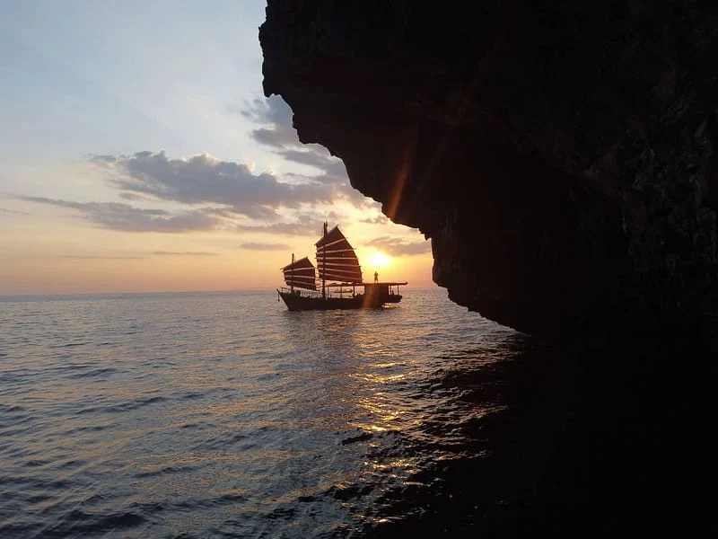 Krabi Sunset Cruises’ red-sailed wooden junk boat beside a limestone cliff at sunset.
